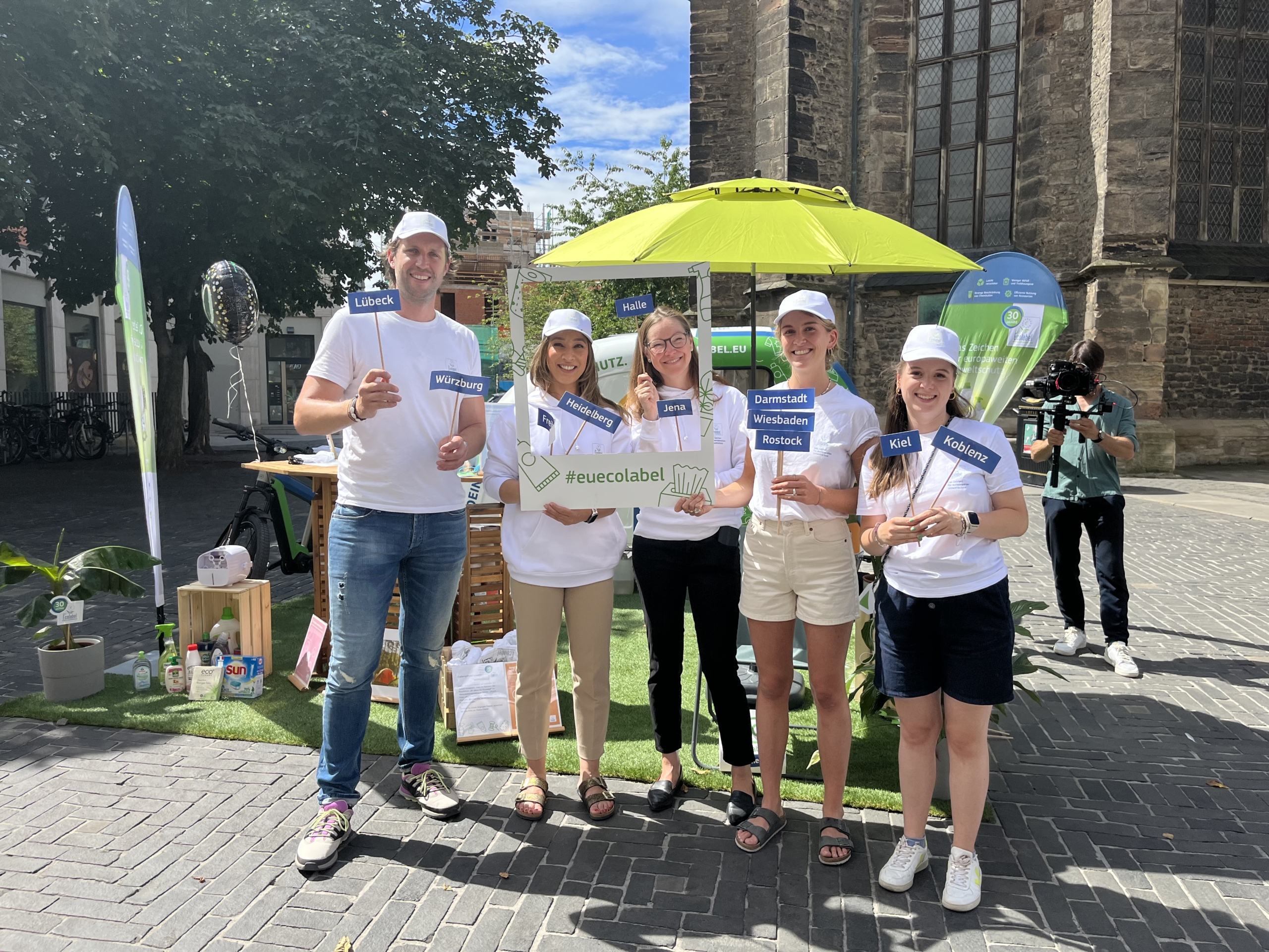 Eine Gruppe von Mitarbeitern steht vor einem aufgebauten Stand von Eu-Ecolabel und hält Städtenamen hoch